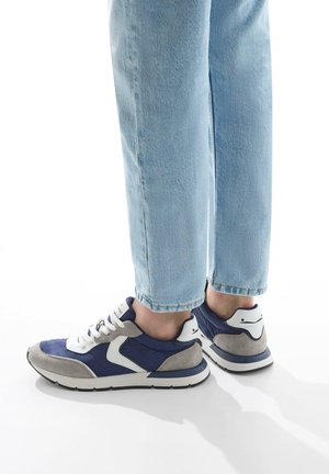 Marineblå og grå træningssko med hvide detaljer, med en struktureret overdel, polstret krave og gummisål. Båret med lyseblå jeans.
