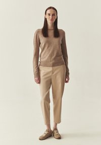 Femme debout, face à l'avant, portant un pull beige, un pantalon court beige clair et des baskets beige sur un fond neutre.