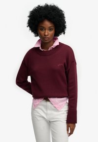 Bordowy sweter z dzianiny z prążkowanymi mankietami, noszony na jasnoróżowej koszuli z kołnierzykiem. Na górnym lewym rogu logo, wykonany z bawełny.