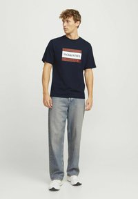 Marinblå t-shirt med en vit och röd rektangulär grafisk design som visar texten "JACK & JONES", kombinerad med ljusblå jeans och vita sneakers.