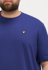 Modrá bavlnená T-shirt s krátkymi rukávmi, s malým žltým vyšívaným logom na hrudi. Hladká textúra, okrúhly výstrih.