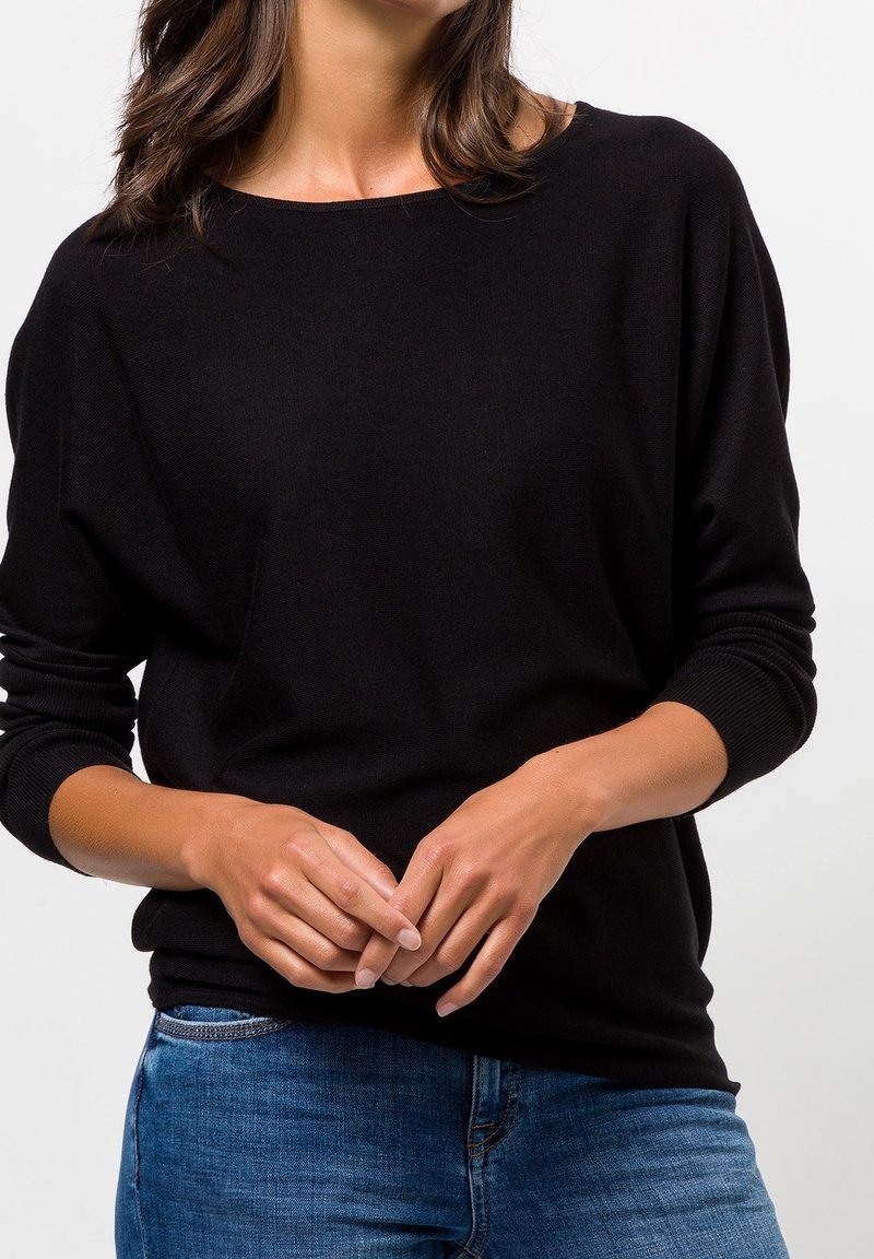 Femme portant un pull noir à manches longues et un jean bleu, les mains doucement jointes devant, sur un fond clair uni.