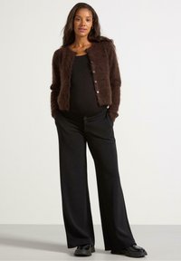 Cardigan marrone delicato sopra una maglietta nera, abbinato a pantaloni neri a gamba larga e scarpe nere; presenta bottoni sulla parte anteriore e una finitura testurizzata.