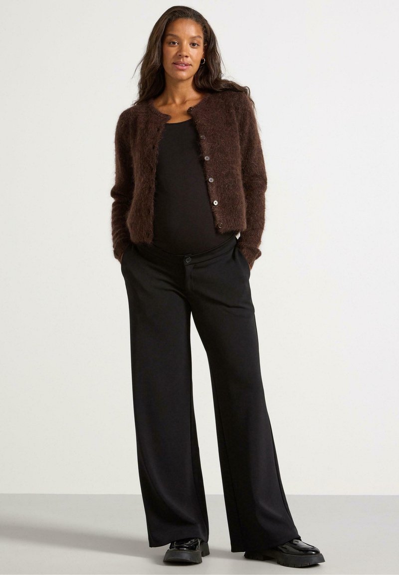 Cardigan marrone delicato sopra una maglietta nera, abbinato a pantaloni neri a gamba larga e scarpe nere; presenta bottoni sulla parte anteriore e una finitura testurizzata.