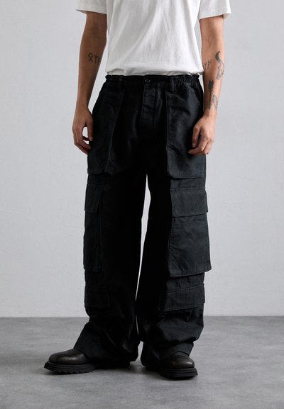 Personne portant un pantalon cargo noir ample avec de grandes poches, une chemise blanche à manches courtes, des bottes noires et des tatouages sur les bras, se tenant sur un sol gris.