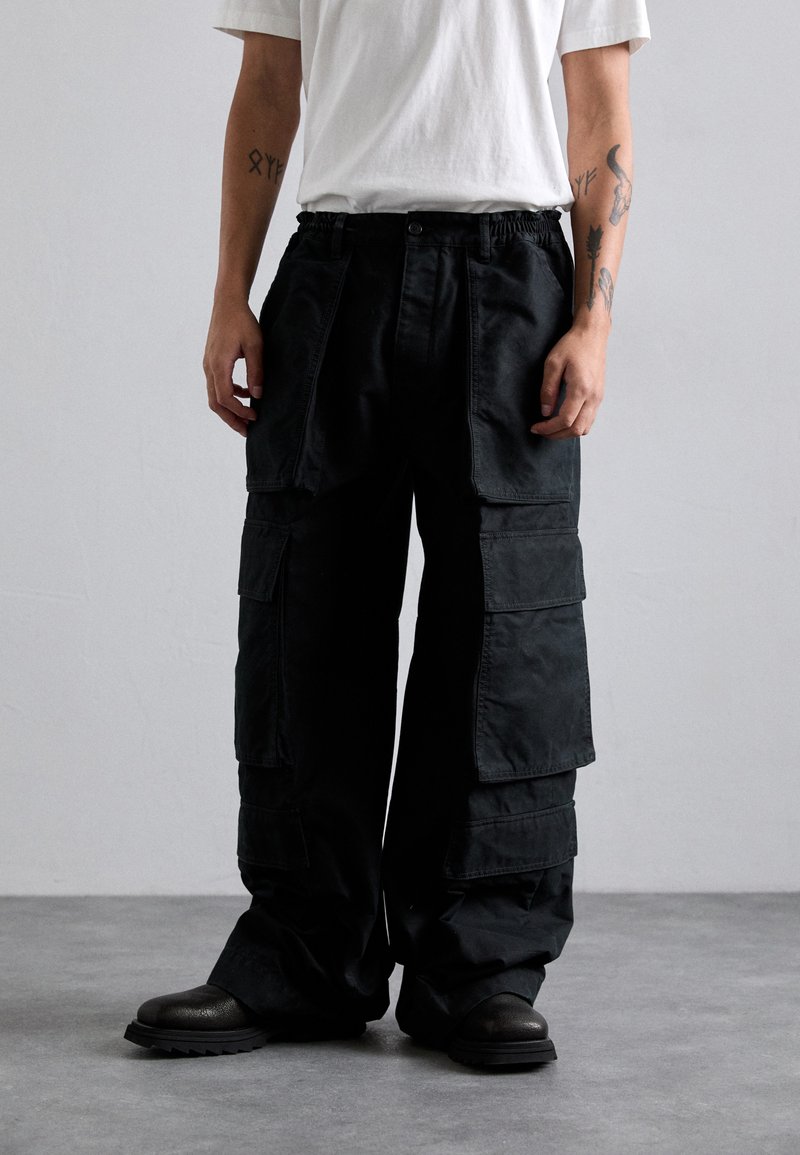 Person wearing loose black cargo pants with large pockets, white short-sleeve shirt, black boots, and arm tattoos standing on gray floor.