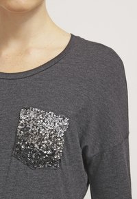 Graues Langarmshirt aus texturiertem Stoff mit einem schimmernden, mit Pailletten besetzten Taschen Design in silbernen Tönen.