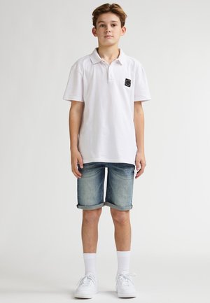 Wit katoenen poloshirt met een logo op de borst, gecombineerd met opgerolde denim shorts. Model draagt witte sneakers en enkelkousen. Neutrale achtergrond.