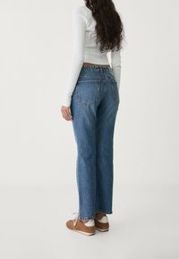 Jeans de denim azul con un corte relajado, que cuentan con cinco bolsillos y una cintura de altura media. Llevados con una camiseta blanca de manga larga y zapatillas marrones.