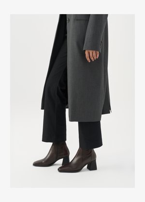 Bottines à talons hauts en cuir marron foncé avec un bout pointu et un talon carré, associées à un pantalon noir et un manteau gris.