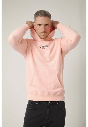 Hombre ajustando la capucha de una sudadera rosa claro con bolsillo frontal y vaqueros negros, fondo neutro.