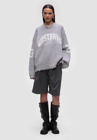 Przesadnie duży szary sweter dzianinowy z prążkowaną teksturą, białymi literami w stylu blokowym i surowym wykończeniem. Połączony z szarymi spódniczkami z plisami i czarnymi wysokimi kozakiem.