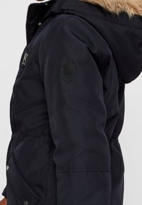 Námornícky modrá parka vyrobená z odolného materiálu, s kapucí lemovanou kožešinou, postranními kapsami a nášivkou na horním rameni.