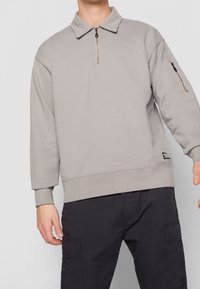 Sudadera de color gris claro con un cuello de cremallera corta, tejido texturizado y un bolsillo con cremallera en la manga izquierda. Puños y dobladillo de canalé.