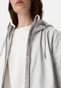 Close-up van een persoon die een lichtgrijze hoodie met trekkoorden en een witte shirt met de tekst "Gemaakt voor dagelijkse avonturen" draagt.