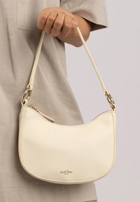 Una mano che tiene una piccola borsa a tracolla di pelle color crema con cerniera dorata e dettagli in metallo, indossata con abbigliamento beige.