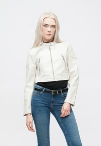 Valge lühike nahktagi külgmiste lukudega, pikad varrukad ja sile tekstuur, koos siniste teksade ja musta topiga.