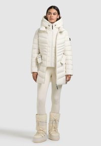 Witte pufferjas met capuchon, verticale stiksels, ritssluiting aan de voorkant en zijzakken. Ggedragen over crèmekleurige legging en witte laarzen met bontvoering.