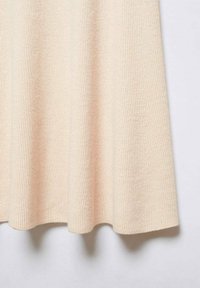 Tissu tricoté côtelé beige suspendu librement, montrant la texture et la chute douce, sur un fond clair uni.