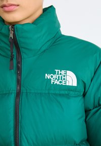 Zaļa izolēta jaka ar augstu apkakli, melnu rāvējslēdzēju un izšūtu balto "The North Face" logo uz krūšu daļas. Gluda tekstūra.