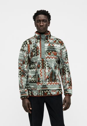 Hombre con jersey polar con estampado de diseños geométricos en gris, negro, blanco y óxido, de pie frente a un fondo blanco.
