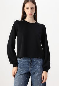 Μαύρη μπλούζα με μακρύ μανίκι και μπαλονέ μανίκια, με διακοσμητικά στοιχεία στους ώμους. Φοριέται με ανοιχτό μπλε τζιν διακοσμημένα με μικρές μεταλλικές τρουκ.