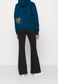 Person som bär en blå Nike-hoodie med guldlogotyp, svarta utsvängda jeans och vita sneakers, vänd bort från kameran mot en vit bakgrund.