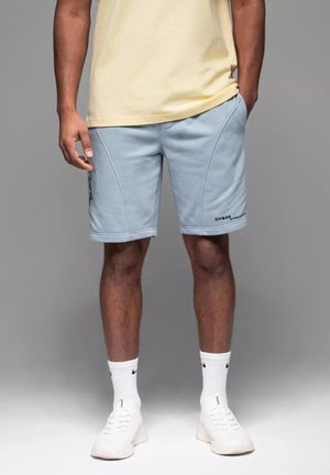 Light blue shorts with an elastic waistband, featuring curved seams and logo detail. Paired with a yellow t-shirt and white sneakers.