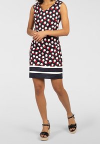 Femme portant une robe noire sans manches à pois rouges et blancs, avec des rayures blanches près de l'ourlet, et des sandales compensées noires, debout sur un sol gris.