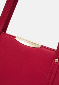 Gros plan d’un sac à main rouge montrant une surface en cuir texturé, une fermeture métallique dorée et une sangle cousue avec un rivet.