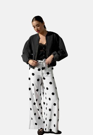 Donna che sistema il cordoncino sui pantaloni bianchi a gamba larga con grandi pois neri, indossando un top in pizzo nero, una giacca bomber nera e scarpe nere.