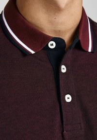 Gros plan d'une personne portant un polo bordeaux avec des boutons blancs et un col avec des rayures blanches et bleu marine.