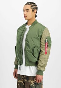 Jachetă bomber verde cu un finisaj mat și manșete striate, având un mânecă de un verde deschis contrastant și un detaliu cu etichetă roșie pe braț.