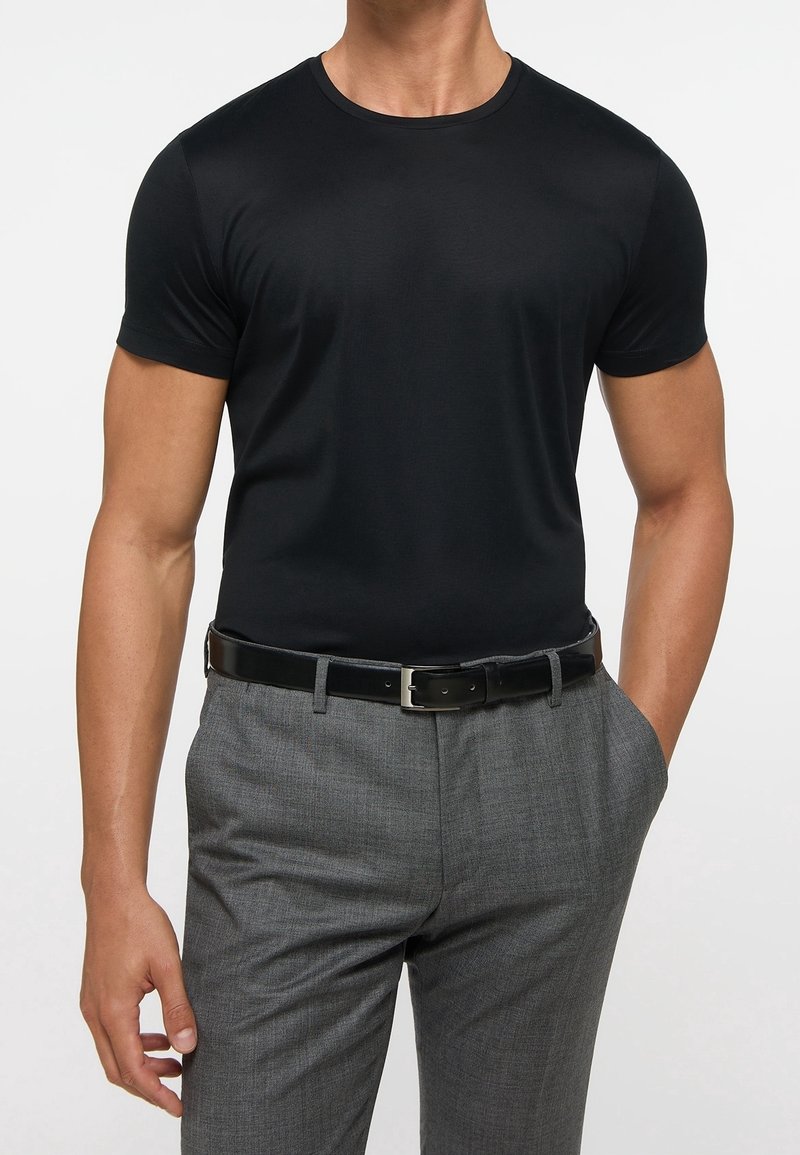 Homme portant un t-shirt noir ajusté à manches courtes, rentré dans un pantalon habillé gris avec une ceinture en cuir noire, main dans la poche.