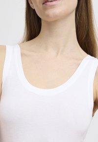 Valge triiksärk, mis on valmistatud sileda ja kerge kangaga, laia kaeluse ja avatud varrukatega disainiga. Keskendutakse ülakehale.