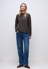 Donna bionda che indossa un maglione marrone scuro, jeans a gamba larga blu e stivaletti in camoscio marroni, in piedi con una mano in tasca.