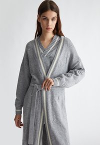 Accappatoio grigio in maglia con scollatura a V, maniche ampie e cintura. Presenta dettagli a righe in un grigio più chiaro e motivi testurizzati.