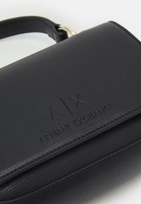Bolso de mano de piel negra con un diseño estructurado, logo en relieve "A|X ARMANI EXCHANGE" y una superficie texturizada sutil.