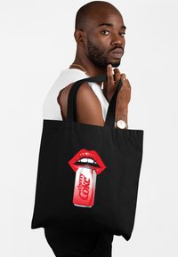 Czarna torba na ramię z grafiką czerwonych ust trzymających puszkę Cherry Coke, z wyrazistymi czerwonymi i białymi detalami. Wykonana z materiału kanwasowego, z solidnymi uchwytami.