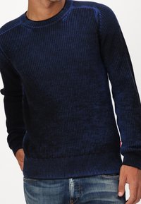 Mörkblå stickad tröja med ribbad struktur och rund halsringning. Har långa ärmar och en något lös passform, kombinerad med denimjeans.