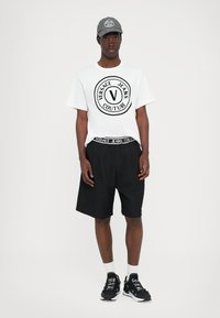 Balta kokvilnas T-kreklu ar melnu Versace Jeans logo, melni šorti ar elastīgu jostasvietu un pelēks cepure; apvienots ar melniem sporta apaviem un baltiem zeķēm.