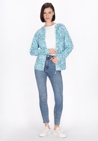 Jachetă cu fermoar de culoare albastru deschis, cu un model floral, purtată peste un top alb și asortată cu blugi albaștri cu talie înaltă și adidași albi.