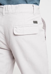 Pantalon beige clair en coton avec une poche arrière, comportant une petite étiquette métallique. Le design comprend des lignes cousues le long de la ceinture.