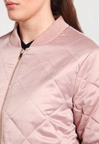 Chaqueta bomber acolchada en suave satén rosa, con cuello acanalado y cremallera brillante, con patrón de diamantes y textura lisa.