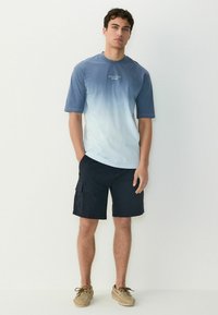 Lys blå gradient t-shirt med kort ærme og logo, parret med navy cargo shorts og brune sko, med en afslappet pasform og casual design.