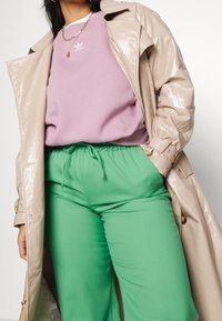 Beige, glänsande trenchcoat över en rosa sweatshirt med vitt logotyp, tillsammans med gröna byxor med dragsko, som visar upp släta texturer och djärva färger.