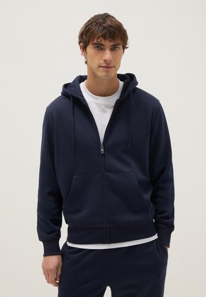 Young man wearing a navy blue zip-up hoodie over a white t-shirt, with hands relaxed, against a plain light background.