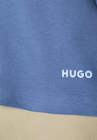 Blauwe katoenen t-shirt met korte mouwen en een rechte zoom. Bevat het witte "HUGO" logo aan de onderzijde links. Lichtbeige stof eronder.