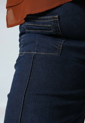 Pantalons en denim bleu foncé avec une texture lisse et des coutures visibles. Présente une poche latérale et une coupe légèrement fuselée.