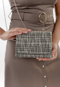 Kobieta trzyma w dłoni torebkę kopertówkę w czarno-białą fakturowaną kratkę z srebrnym łańcuchem, ubrana w taupe sukienkę z paskiem i złotymi guzikami.